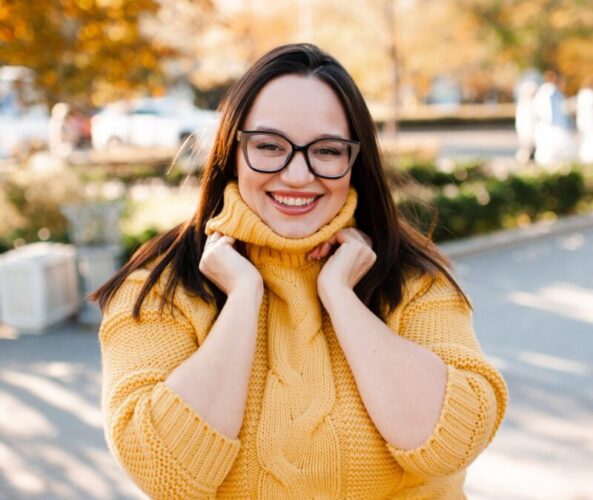 Smiling mature woman 35-40 year old wearing eye glasses and knitted yellow sweater over autumn city background close up. Fall season. Happiness.